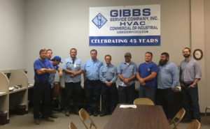 A group of ten men in blue work shirts stand in a row inside an office. Behind them, a banner reads GIBBS Service Company, Inc. HVAC Celebrating 45 Years. There are desks, chairs, and a wall clock in the room.